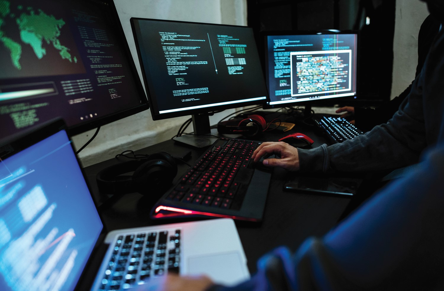 A workspace with multiple monitors displaying code, a laptop, and a person typing on a mechanical keyboard.