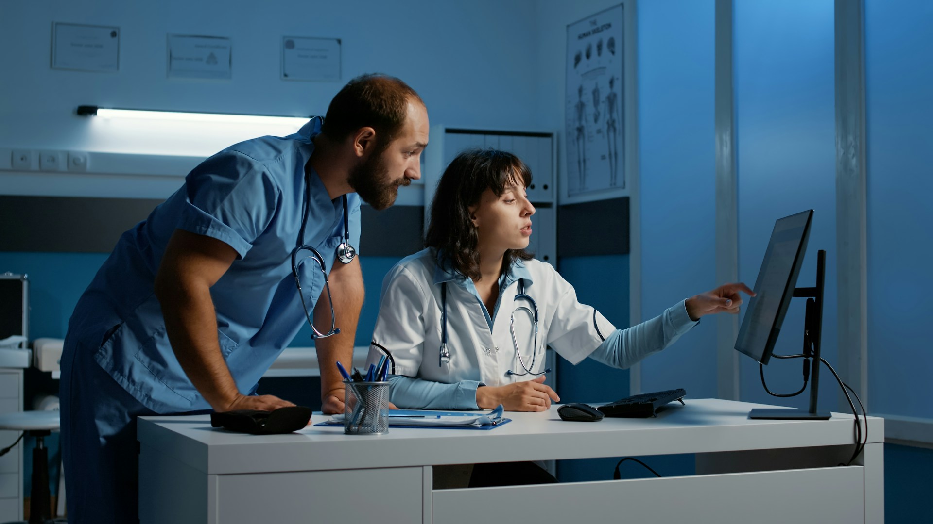 A doctor and nurse discuss patient data on a computer in a medical office, surrounded by medical charts and equipment.