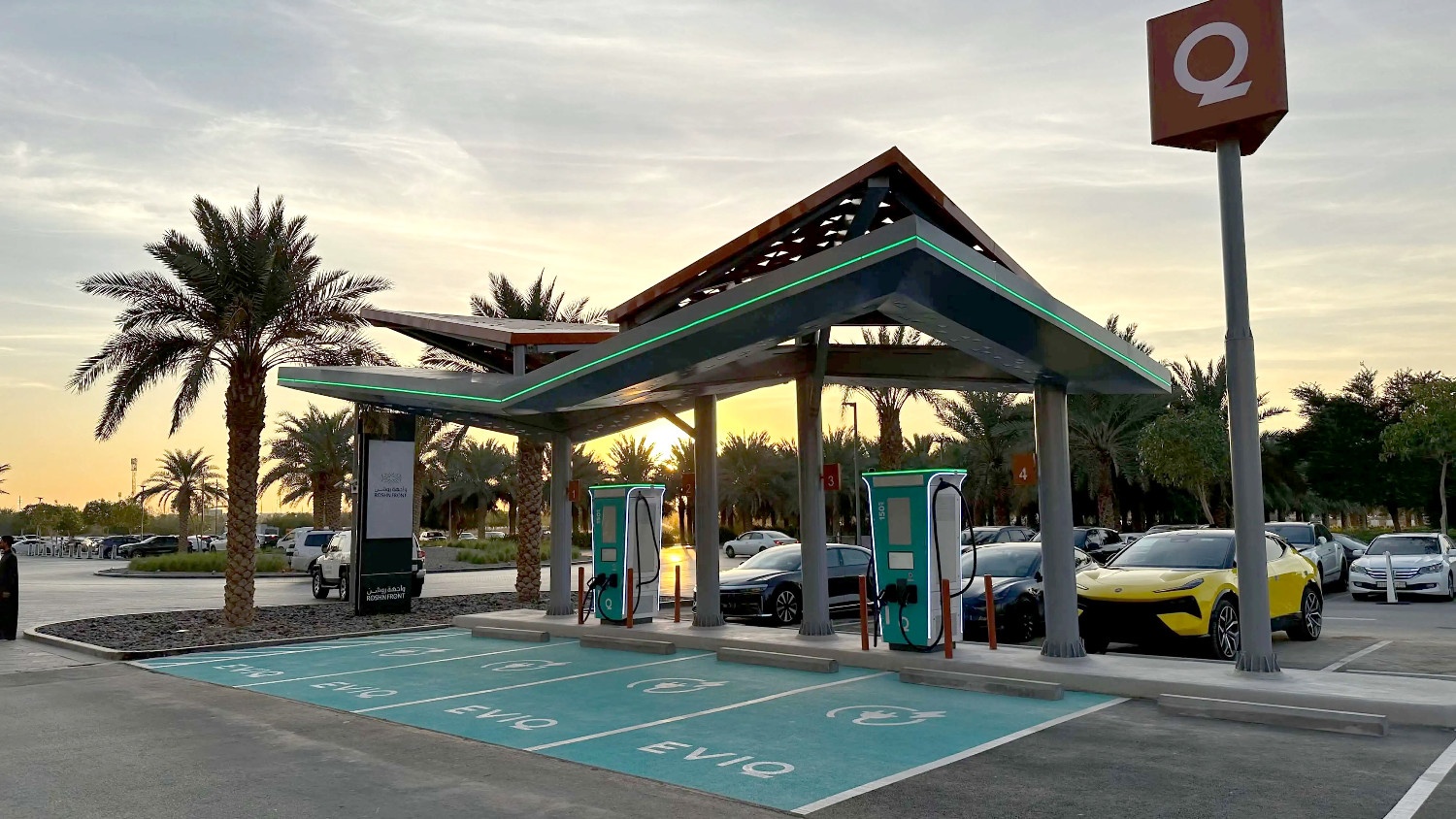 Electric vehicle charging station with multiple chargers under a modern canopy, palm trees, and parked cars at sunset.